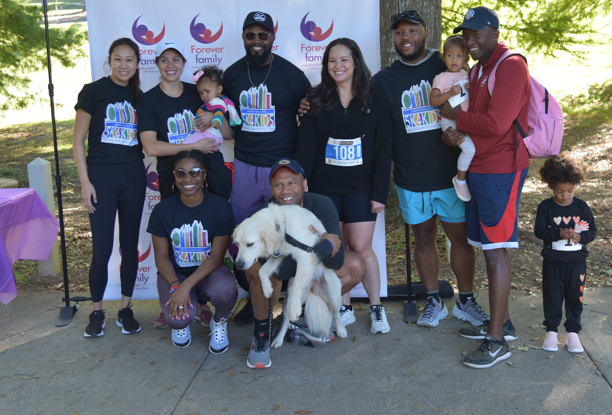 Diverse group of 8 adults, 2 toddlers, and a white dog posing outside in front of the Forever Family 5k run banner.